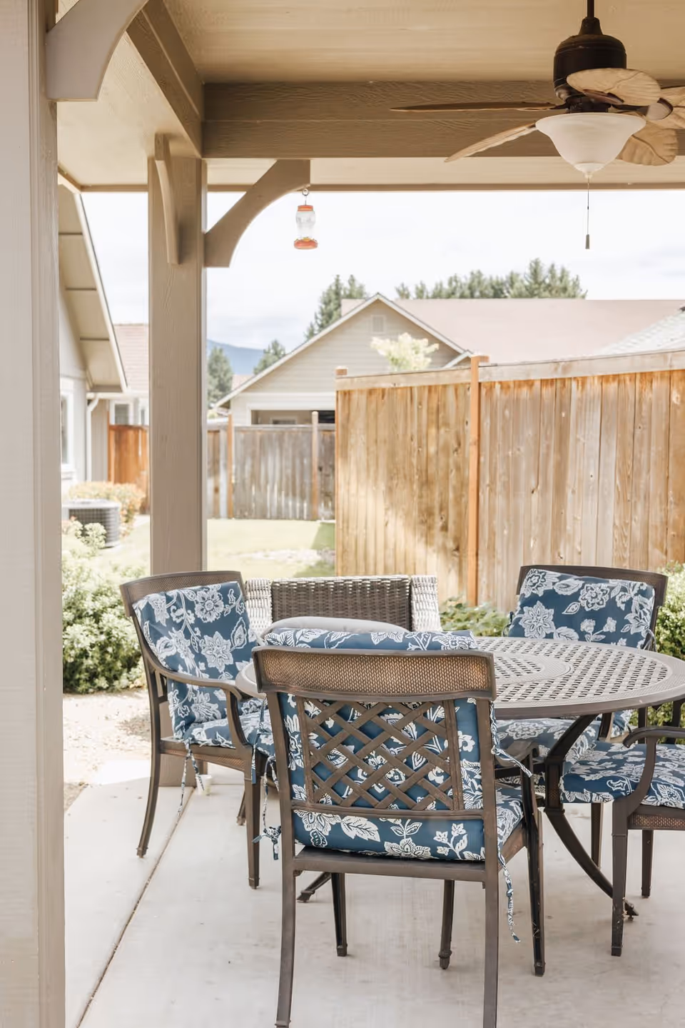 Covered outdoor patio area with a round metal table and four chairs with blue and white floral cushions. A ceiling fan with wooden blades hangs above the table. In the background, there is a wooden fence and neighboring houses with trees visible.