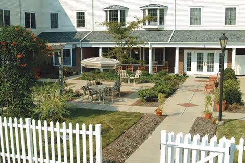Courtyard of an assisted living facility featuring patio tables and an umbrella, landscaped walkways, a white picket fence, and the building entrance.