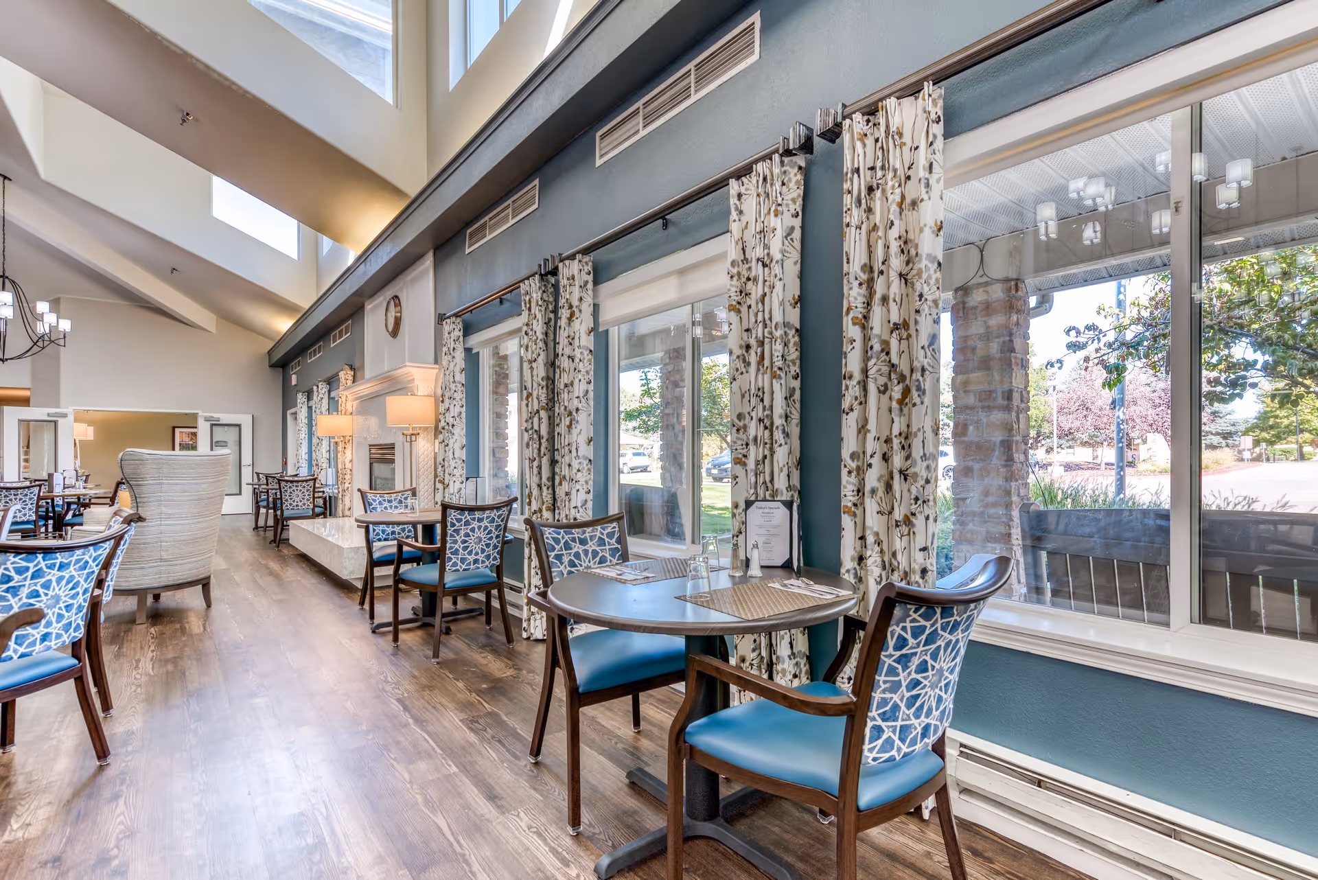 A bright dining area in a senior living facility with multiple tables and chairs arranged along large windows with floral curtains. The room features high ceilings with skylights, wooden flooring, and a fireplace with lamps on either side. Outside the windows, trees and a street are visible.