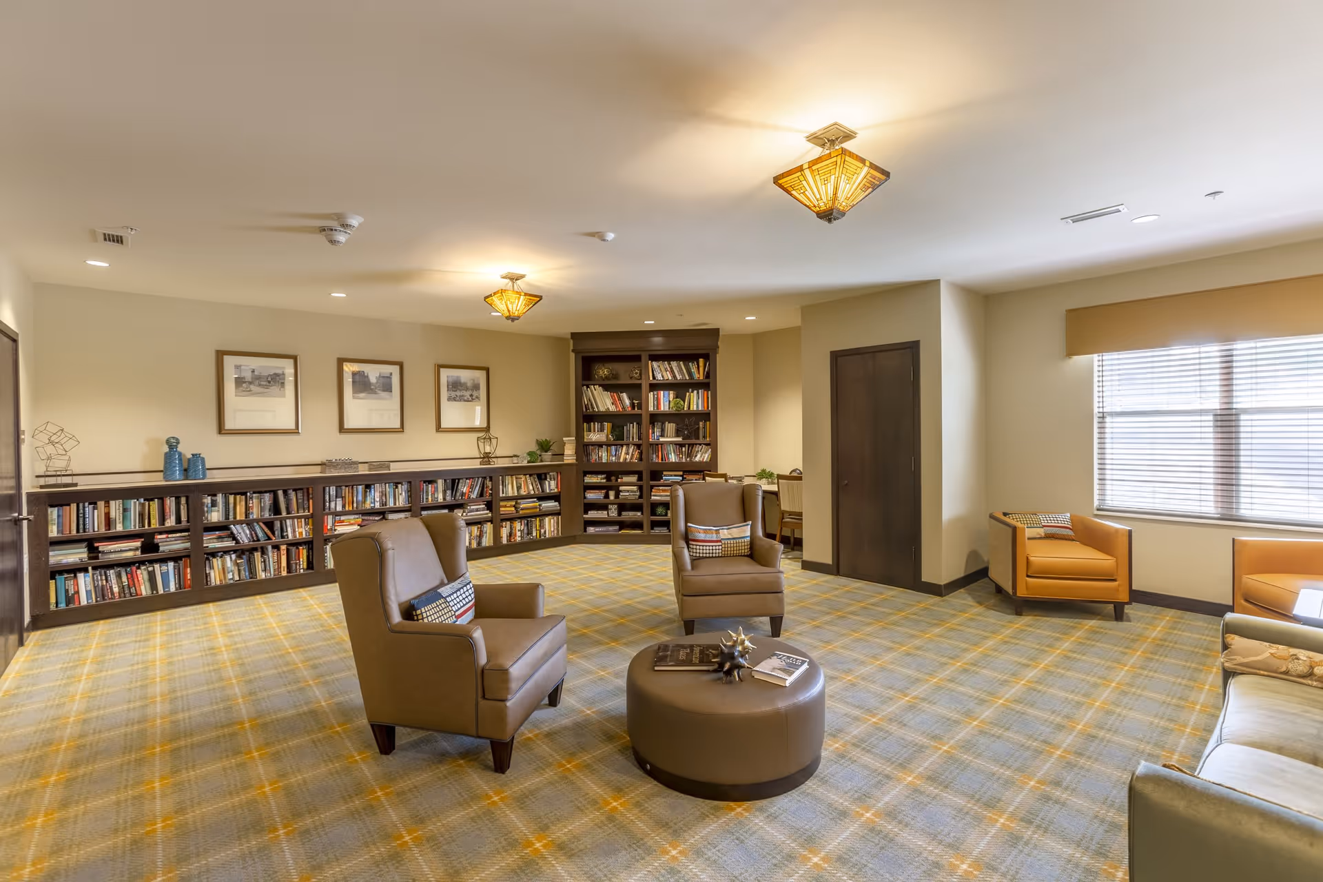 A spacious and well-lit living room area with a patterned carpet, two brown leather armchairs with decorative pillows, a round ottoman with books and decor on top, a large bookshelf filled with books along the back wall, and a window with blinds letting in natural light. The walls are light-colored and there are framed pictures hanging above the bookshelf.