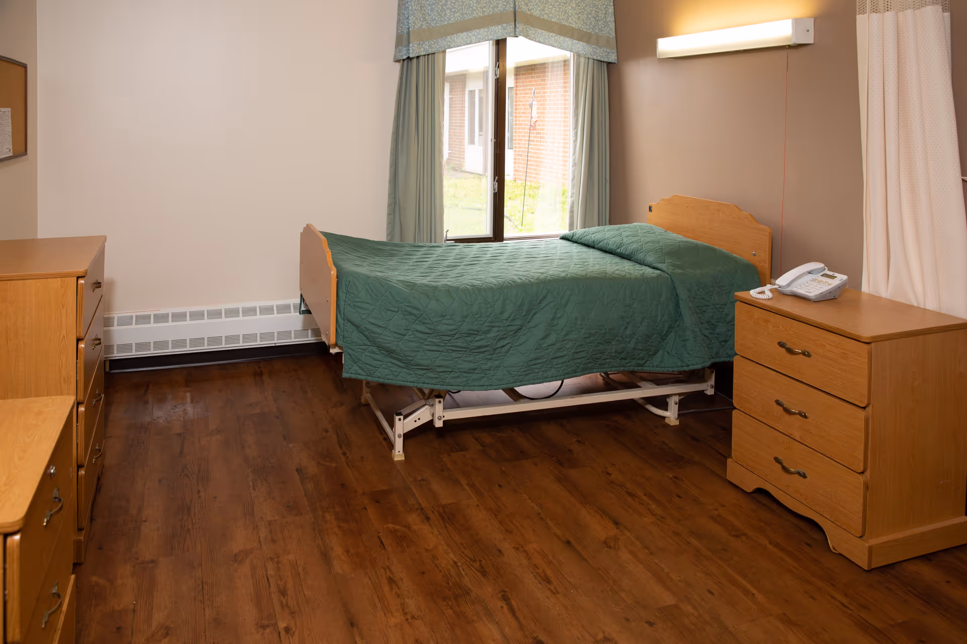 A simple bedroom in a senior living facility with a single bed covered with a green quilt, wooden dressers on either side, a telephone on one dresser, a window with curtains, and wooden flooring.