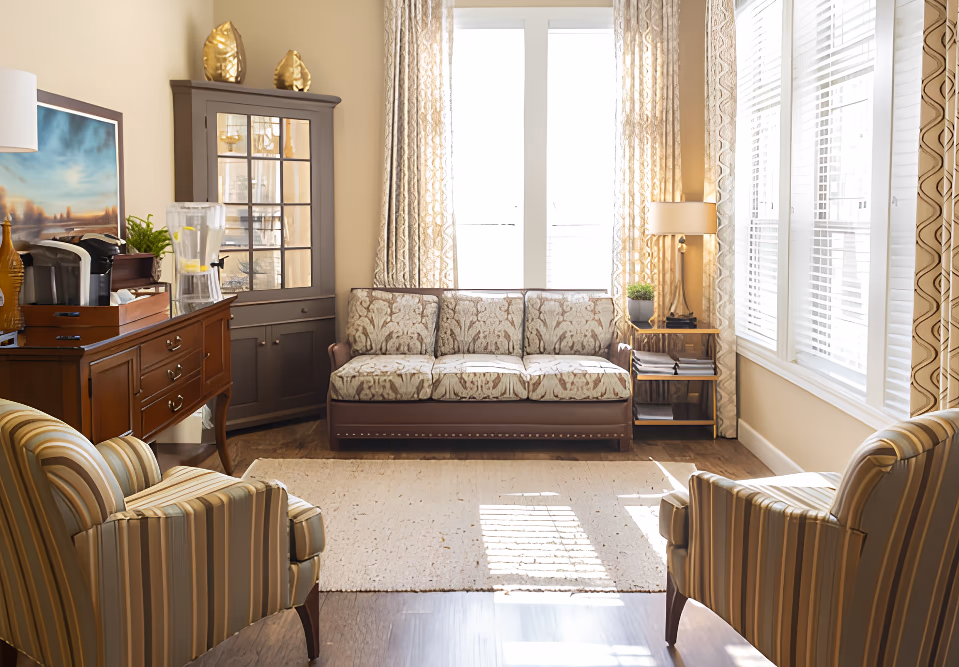 A bright and cozy living room with a patterned three-seater sofa against a window with sheer curtains. Two striped armchairs face the sofa across a light-colored rug on a wooden floor. To the left, there is a wooden sideboard with a coffee maker and a water dispenser, and a corner cabinet with decorative items on top. A floor lamp and a small side table with books and a plant are on the right side near the window.