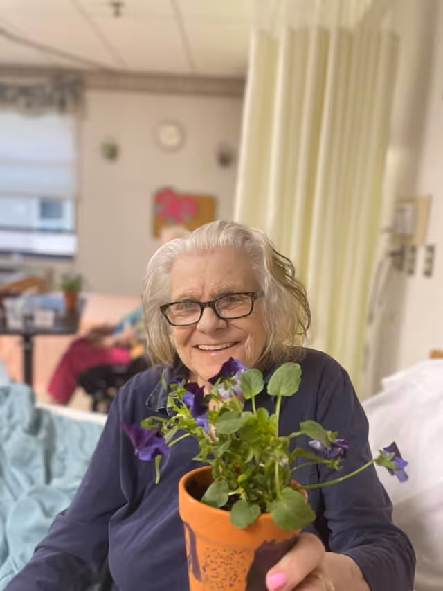 An elderly woman with white hair and glasses is sitting on a bed in a room, smiling and holding a small potted plant with purple flowers. The background shows a curtain, a clock on the wall, and another person sitting at a table.