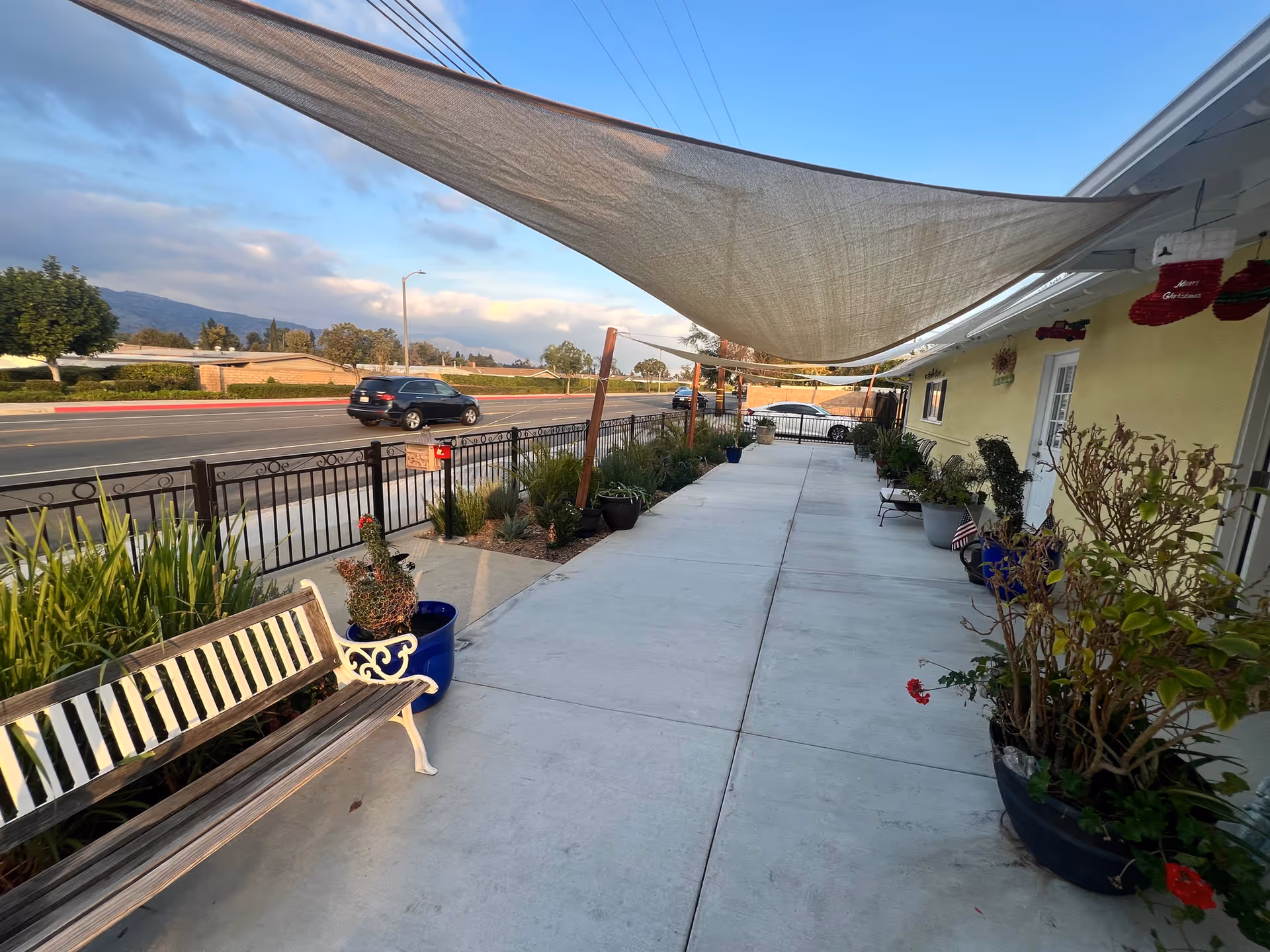 Covered front walkway of a senior residence with benches, potted plants, a shade canopy, and a metal fence along the street.