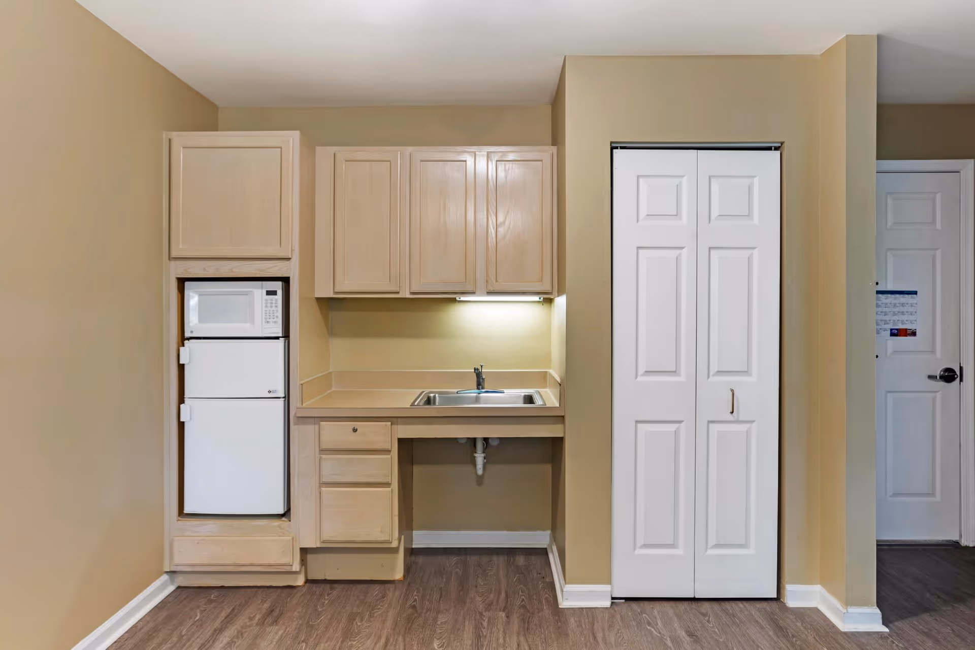 Small kitchenette area with light wood cabinets, a white microwave and mini refrigerator stacked on the left, a stainless steel sink with a faucet under a cabinet with under-cabinet lighting, and a white bi-fold door closet on the right. The walls are beige and the floor is wood laminate.