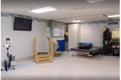 A physical therapy or rehabilitation room with exercise equipment including a stationary bike, parallel bars with wooden steps, a treatment table, and various therapy tools and mats against the walls. A wall-mounted TV is visible on the left side.