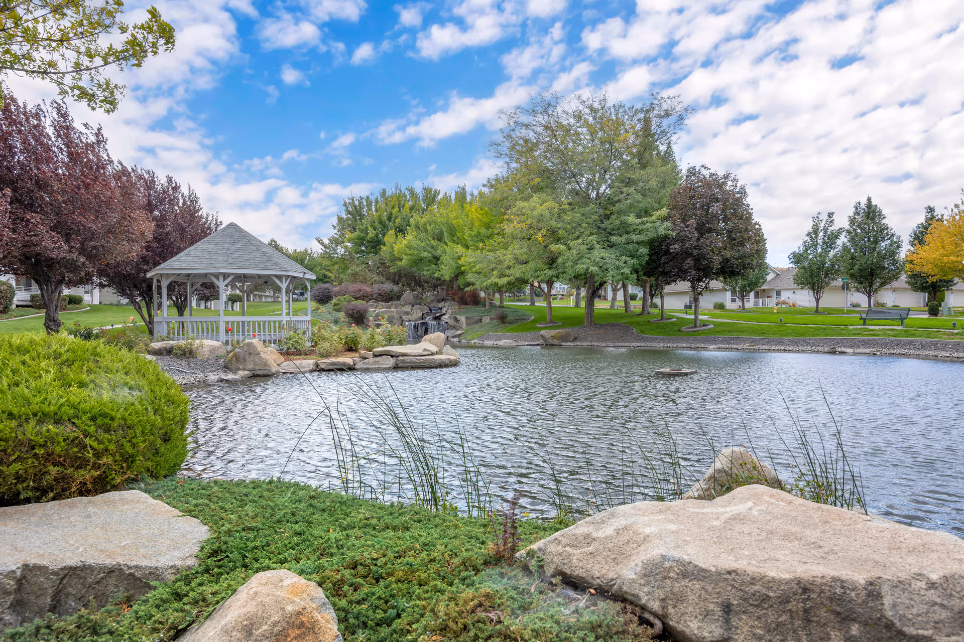 A serene outdoor scene at Brookdale Canyon Lakes featuring a small pond surrounded by rocks and greenery. There is a white gazebo on the left side near the water, with various trees and bushes around the pond. The sky is partly cloudy with patches of blue.