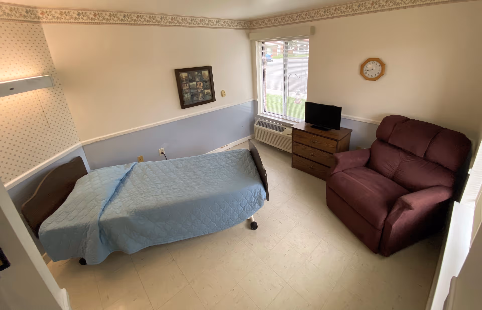 A small bedroom in a senior living facility with a single bed covered with a light blue quilt, a maroon recliner chair, a wooden dresser with a small flat-screen TV on top, a window with blinds, a wall clock, and a framed picture on the wall.