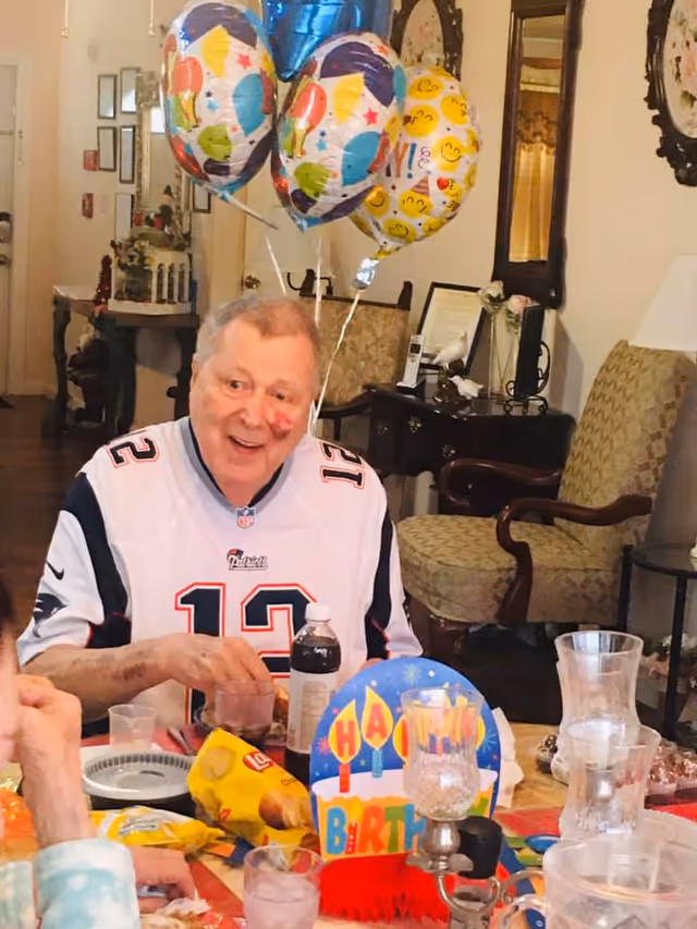 An elderly man wearing a New England Patriots jersey is sitting at a table decorated for a birthday celebration with balloons and a colorful Happy Birthday sign. The table has snacks, drinks, and party items. The background shows a cozy room with chairs, a mirror, and decorative items.