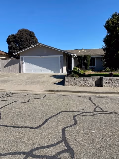 Single-story residential house with a two-car garage, a driveway, and a small front yard with shrubs and trees under a clear blue sky.