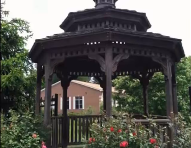 A wooden octagonal gazebo in a garden with flowering bushes and a residential building visible behind it.