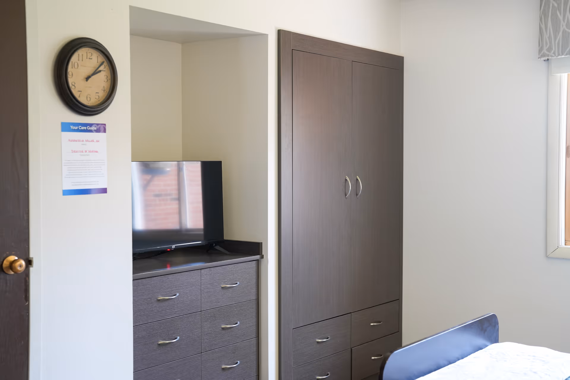 A bedroom corner with a wall clock, a mounted TV on a dark wooden dresser, a matching wardrobe, and a window letting in natural light. Part of a bed with white bedding is visible in the foreground.