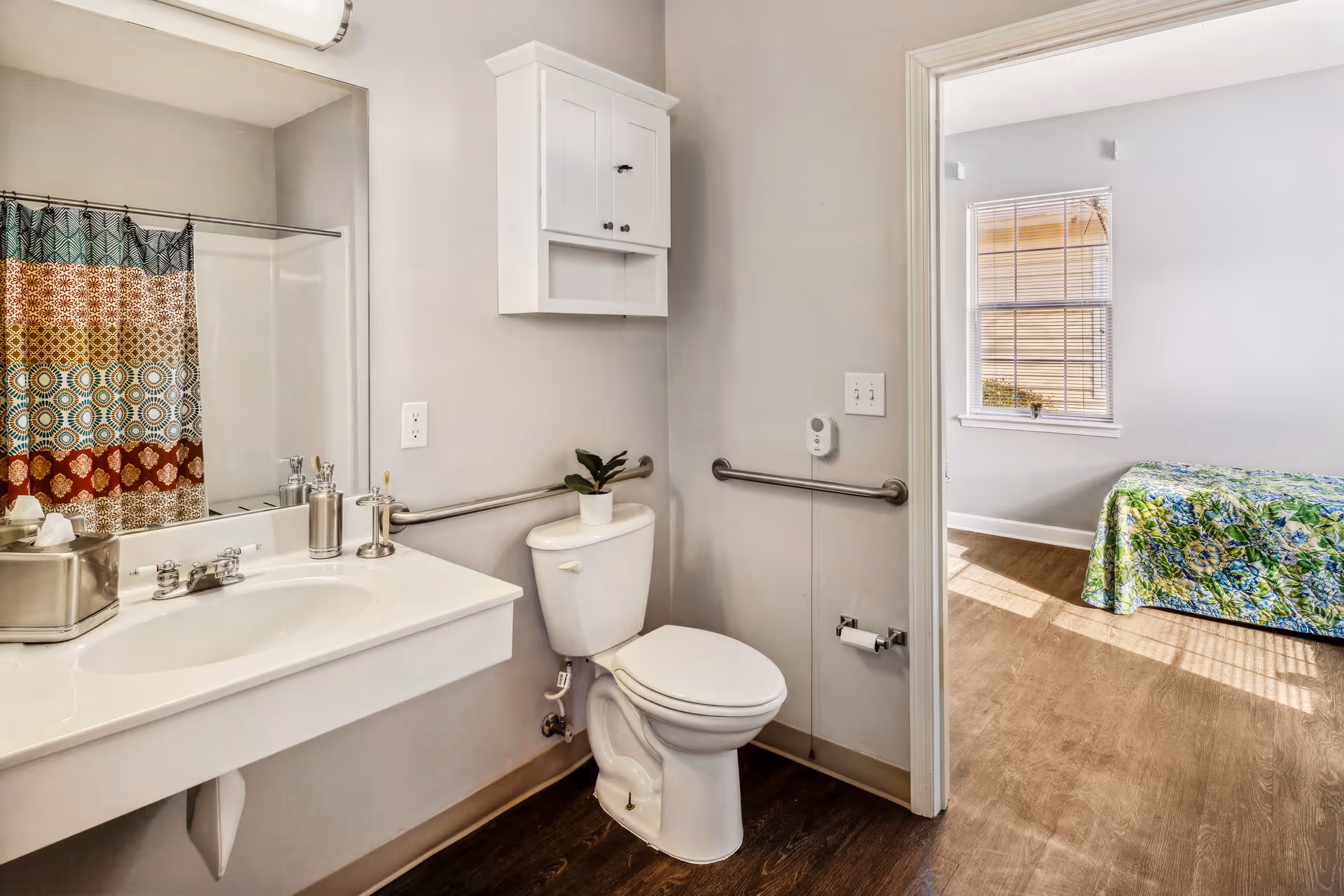 A bathroom with a white sink and countertop, a toilet with grab bars on both sides, a colorful patterned shower curtain, and a white wall cabinet above the toilet. The bathroom opens into a bedroom with a window and a bed covered with a green and blue patterned bedspread.