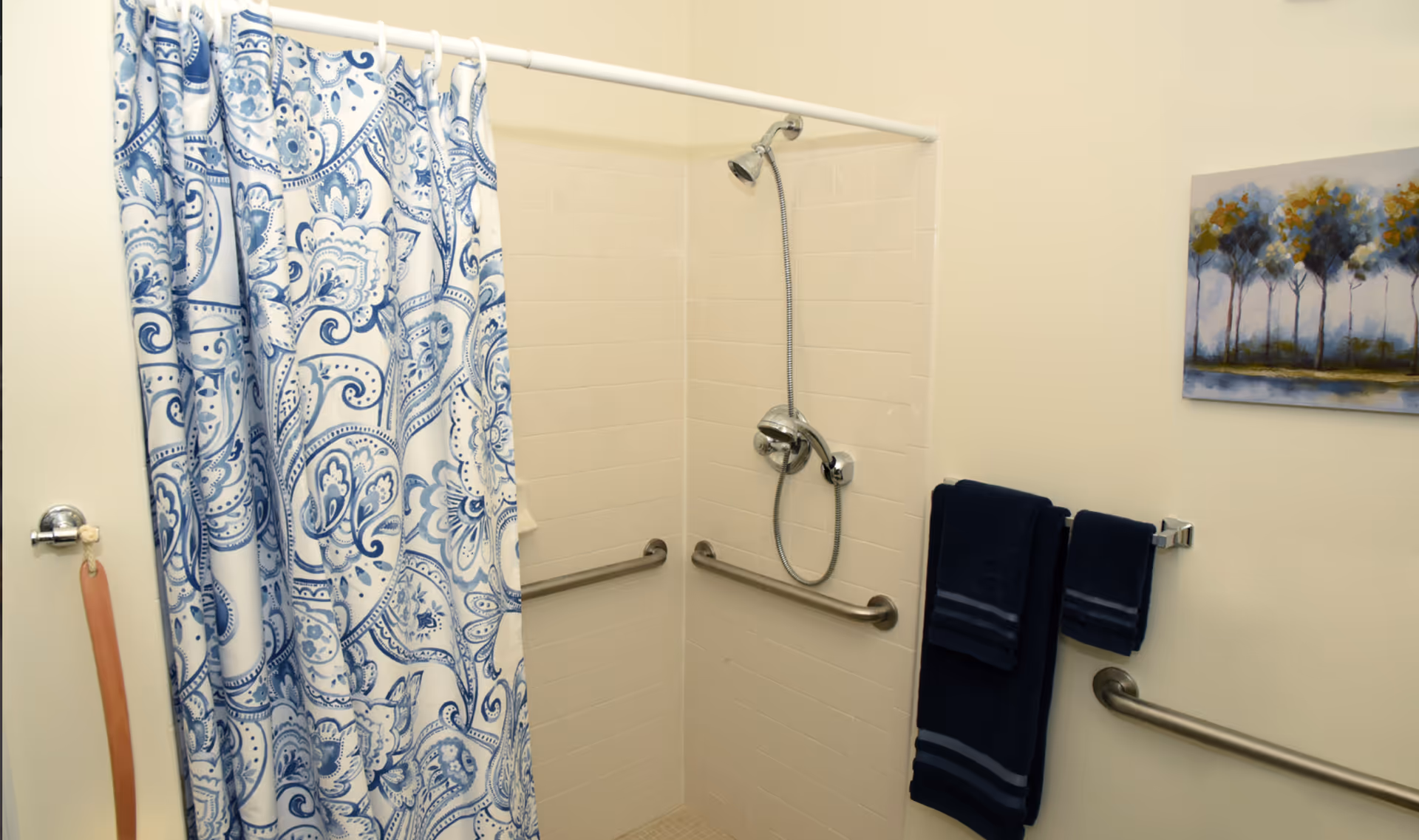 A bathroom shower area with white tiled walls and a blue and white patterned shower curtain. There are two metal grab bars installed on the walls inside the shower and one outside. Two dark blue towels hang on a towel rack next to the shower. A painting of trees is hung on the wall above the towels.