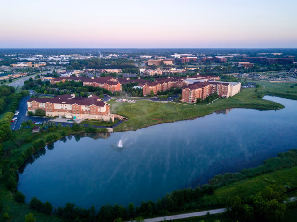 Aerial view of a large senior living facility named Sedgebrook, featuring multiple connected buildings with red roofs situated next to a pond with a water fountain. Surrounding the facility are green lawns, trees, and a nearby road with additional buildings in the background under a clear sky at dusk.