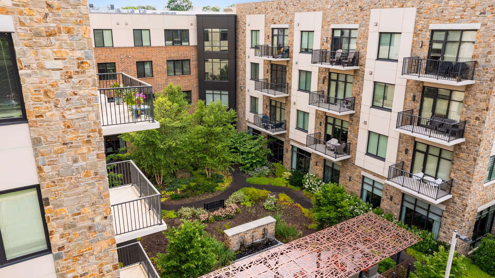 View of an outdoor courtyard at Eagleview Landing, surrounded by multi-story residential buildings with balconies. The courtyard features green trees, shrubs, a walking path, and a decorative water feature with a metal pergola structure.
