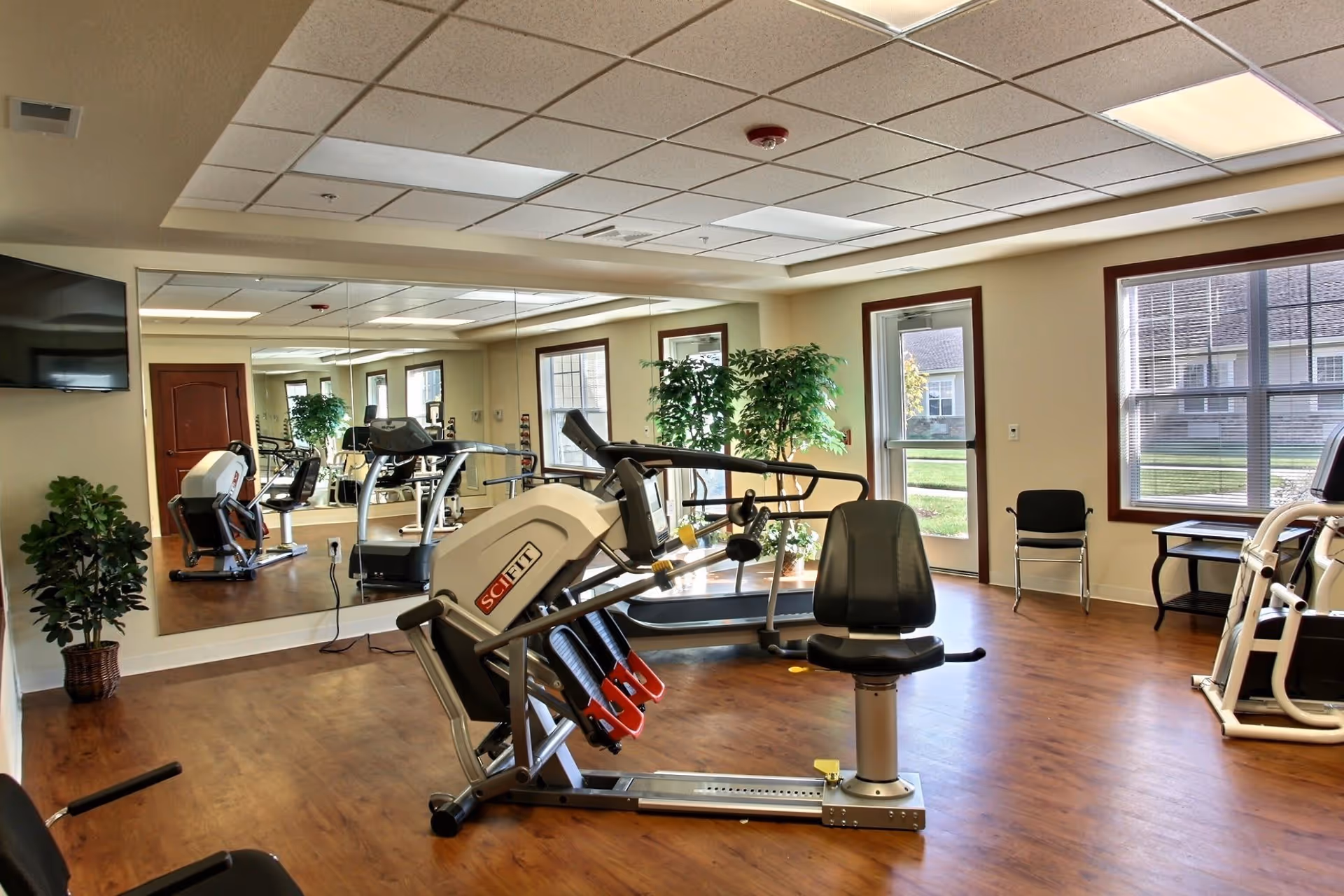 A bright fitness room with exercise machines, a large wall mirror, TV, windows, and wood flooring.