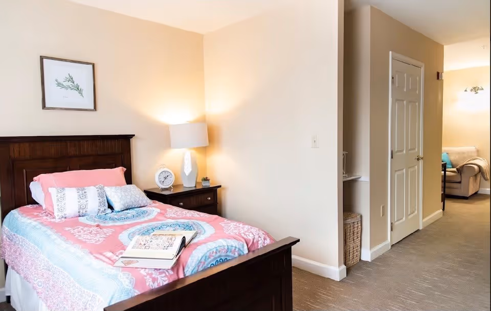 A cozy bedroom with a wooden bed frame and a colorful pink and blue patterned bedspread. There are several pillows on the bed and an open book resting on the bedspread. A nightstand beside the bed holds a white table lamp, a round clock, and a small plant. The room has beige walls and carpeted flooring. In the background, there is a hallway leading to another room with a beige armchair and a small table with a lamp.