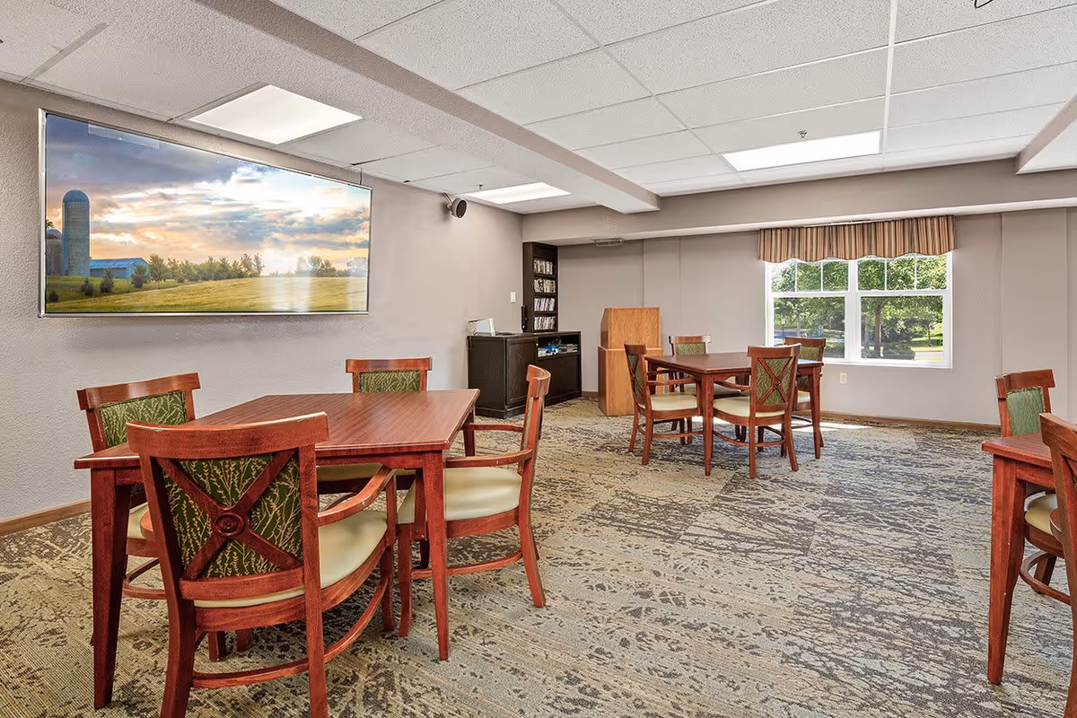 A well-lit room with multiple wooden tables and chairs arranged for seating. The chairs have green patterned upholstery. A large window with a striped valance allows natural light to enter, showing greenery outside. On one wall, there is a large framed picture of a rural landscape with a barn and silo. The room has a carpeted floor with a patterned design and a ceiling with recessed lighting panels.