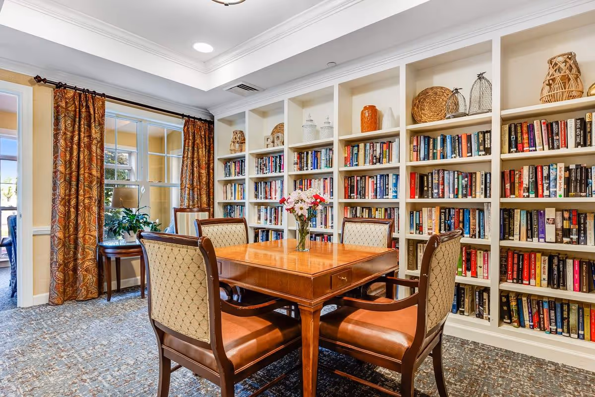 A cozy reading or meeting area in a senior living facility featuring a wooden table with four upholstered chairs. Behind the table is a large built-in bookshelf filled with books and decorative items. The room has patterned curtains on the windows and a small side table with a plant near the window.