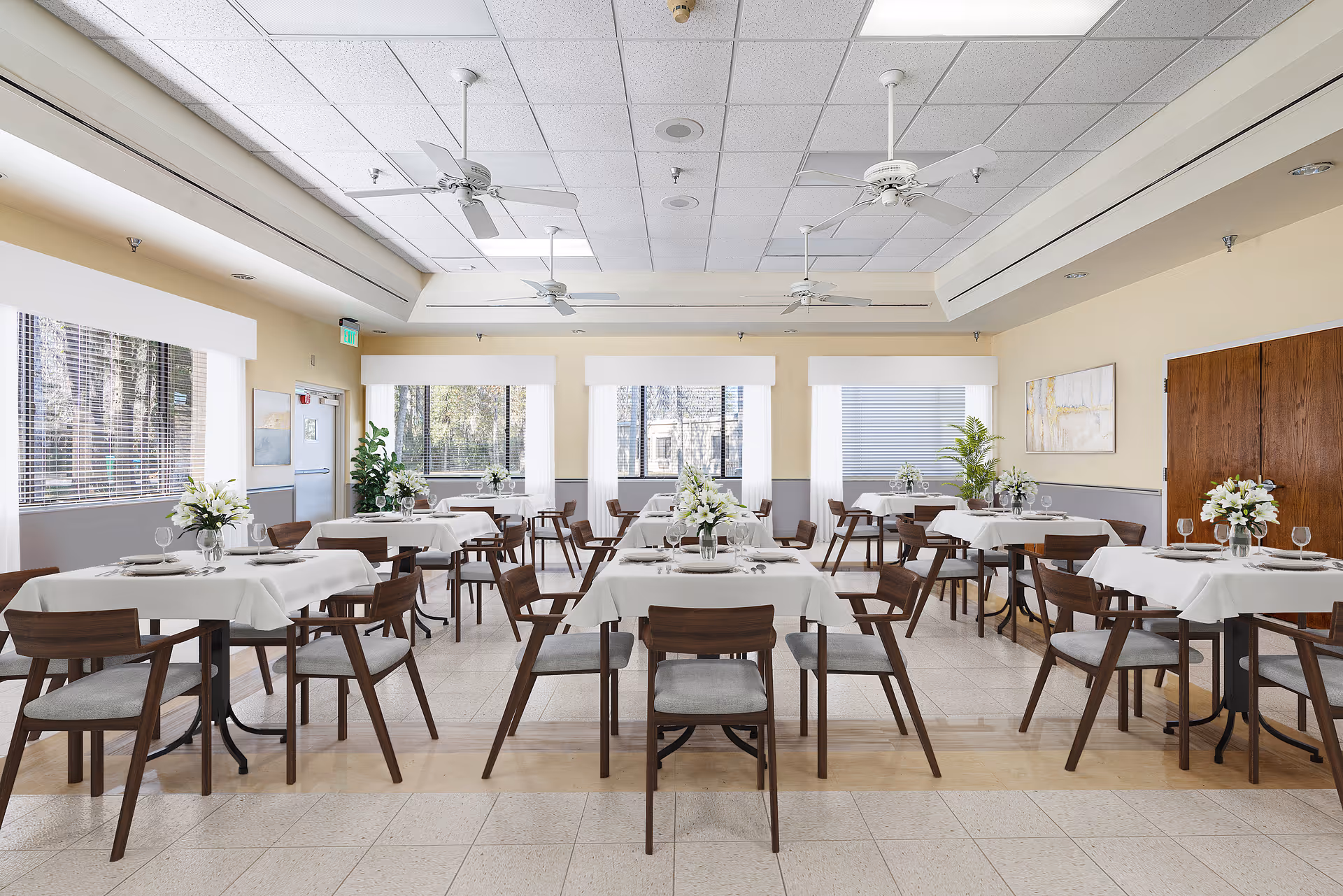 A bright and spacious dining room with multiple tables covered in white tablecloths, each set with plates, glasses, and floral centerpieces. The room has large windows with blinds, ceiling fans, and wooden chairs with gray cushions arranged around the tables.