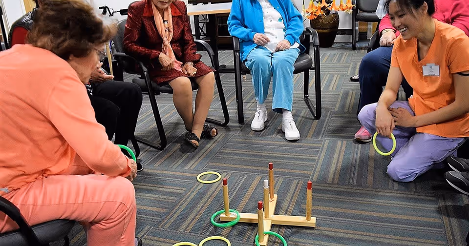 A group of elderly people and a caregiver sitting in a circle playing a ring toss game indoors on a carpeted floor. The caregiver is smiling and holding a ring, while the elderly participants are engaged in the activity.