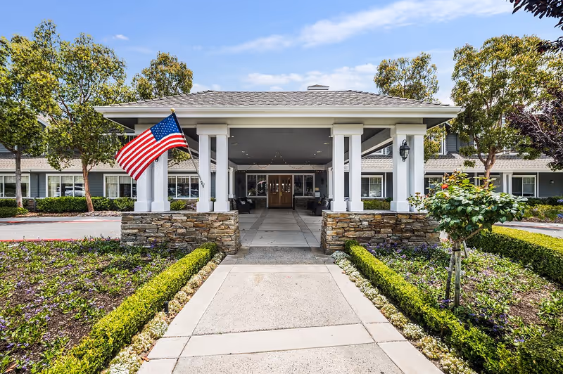Front entrance of a senior living facility with a covered driveway supported by white columns and stone bases, an American flag mounted on the left side, landscaped flower beds and bushes on either side of the walkway, and a building with multiple windows in the background under a partly cloudy sky.
