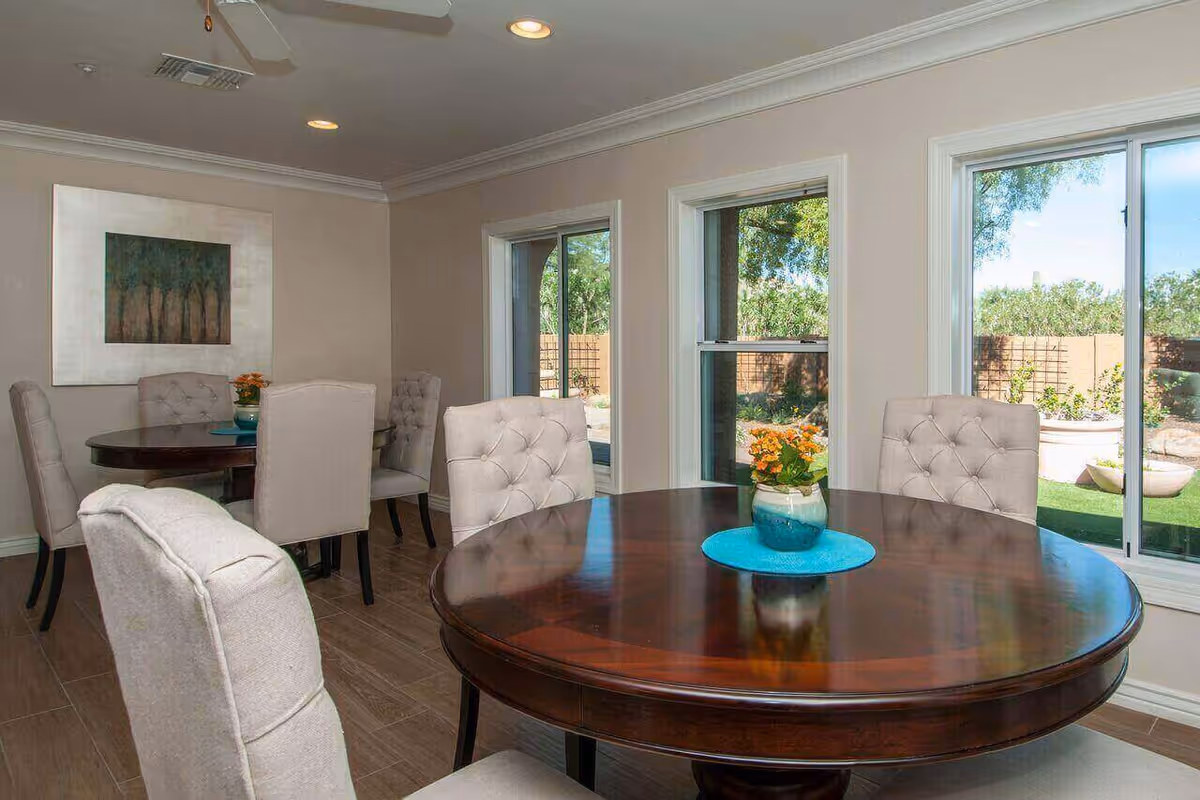 Bright dining room with round wooden tables, upholstered tufted chairs, and large windows looking out to a garden.