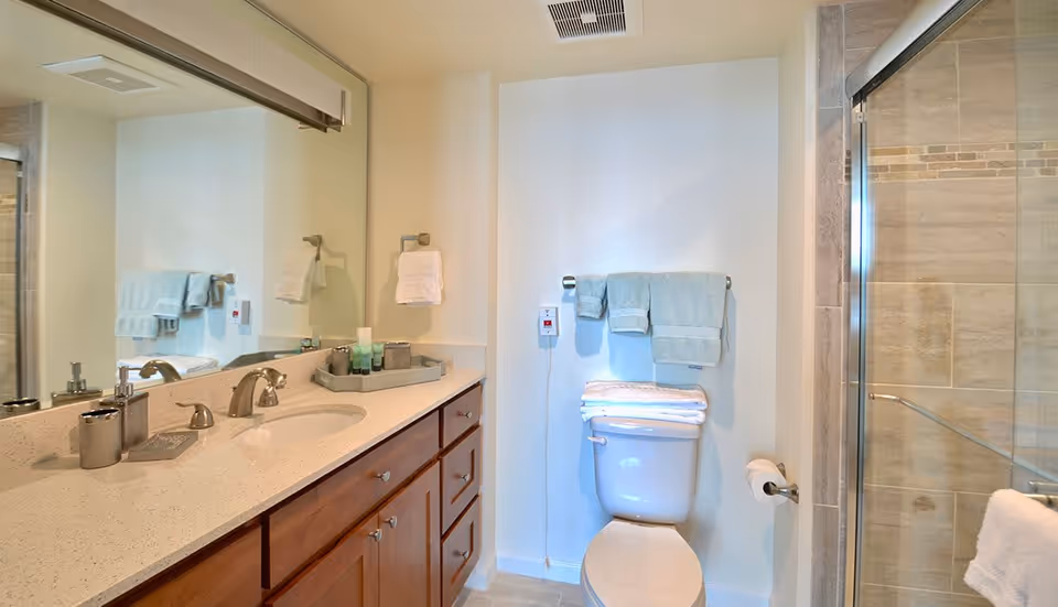 Well-lit bathroom featuring a long vanity with sink and mirror, a toilet with folded towels, and a glass-enclosed shower.
