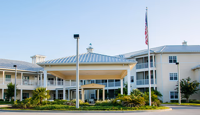 Front entrance of a multi-story senior living building with a covered porte-cochere, flagpole, and landscaped circular drive.