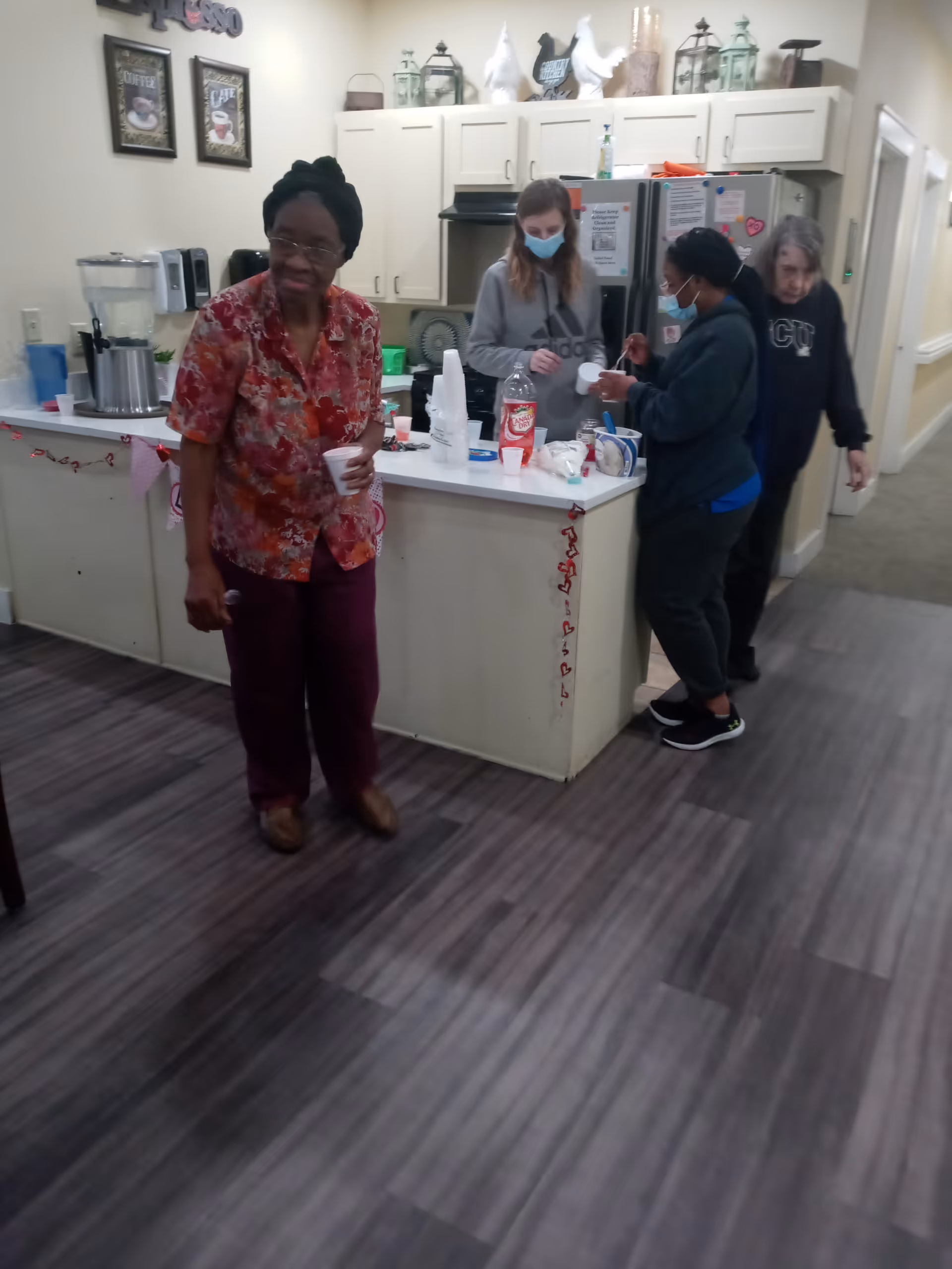 Four people in a kitchen area of a senior living facility. One woman in a red floral shirt is holding a cup and spoon, standing near the camera. Two women wearing masks are preparing drinks at the kitchen counter, which has cups, a bottle of soda, and other items. Another woman is walking in the hallway beside the kitchen. The kitchen has white cabinets, a refrigerator, and decorative items on top of the cabinets.