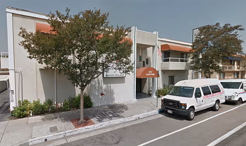 Exterior view of Glen Park at Valley Village facility showing a two-story building with white walls, orange awnings, and an entrance with an orange canopy. Two white vans are parked on the street in front of the building, and there are trees and shrubs along the sidewalk.