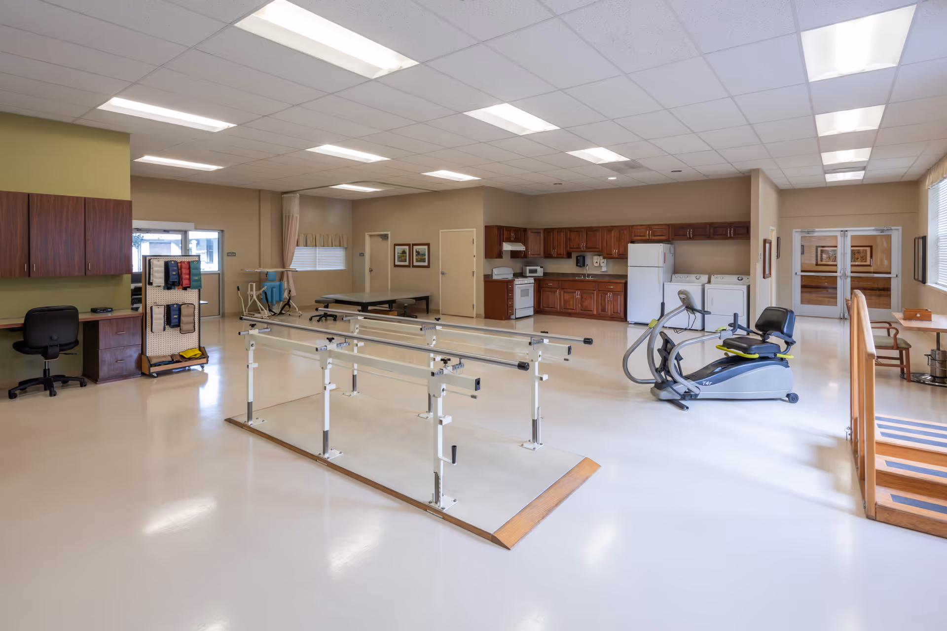 Spacious rehabilitation therapy room with parallel walking bars, exercise bike, practice stairs and a kitchenette area.