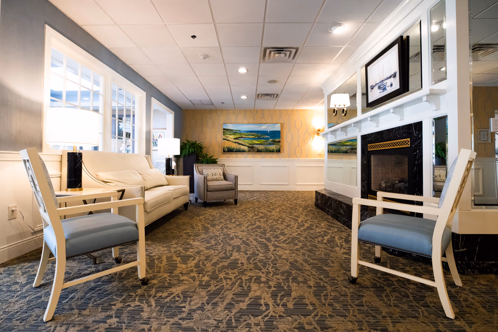 A cozy living room area in The Elms facility featuring a beige sofa, two white chairs with blue cushions, a patterned carpet, a fireplace with a black marble surround, a framed picture above the fireplace, and a landscape painting on the far wall. The room is well-lit with table lamps and wall sconces, and has a mix of light blue and yellow walls with white wainscoting.