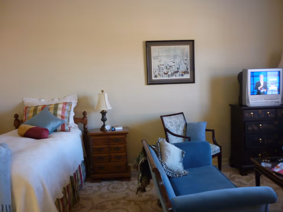 Cozy bedroom with a single bed, nightstand and lamp, blue settee and chairs, and a television on a dresser.