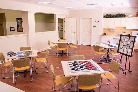 A bright activity room with several tables and chairs arranged for games and socializing. One table has a large checkers board, another has a partially completed puzzle, and a standing board with a crossword puzzle is visible. The room has wood flooring, light-colored walls, and a kitchenette area with cabinets and a countertop in the background.