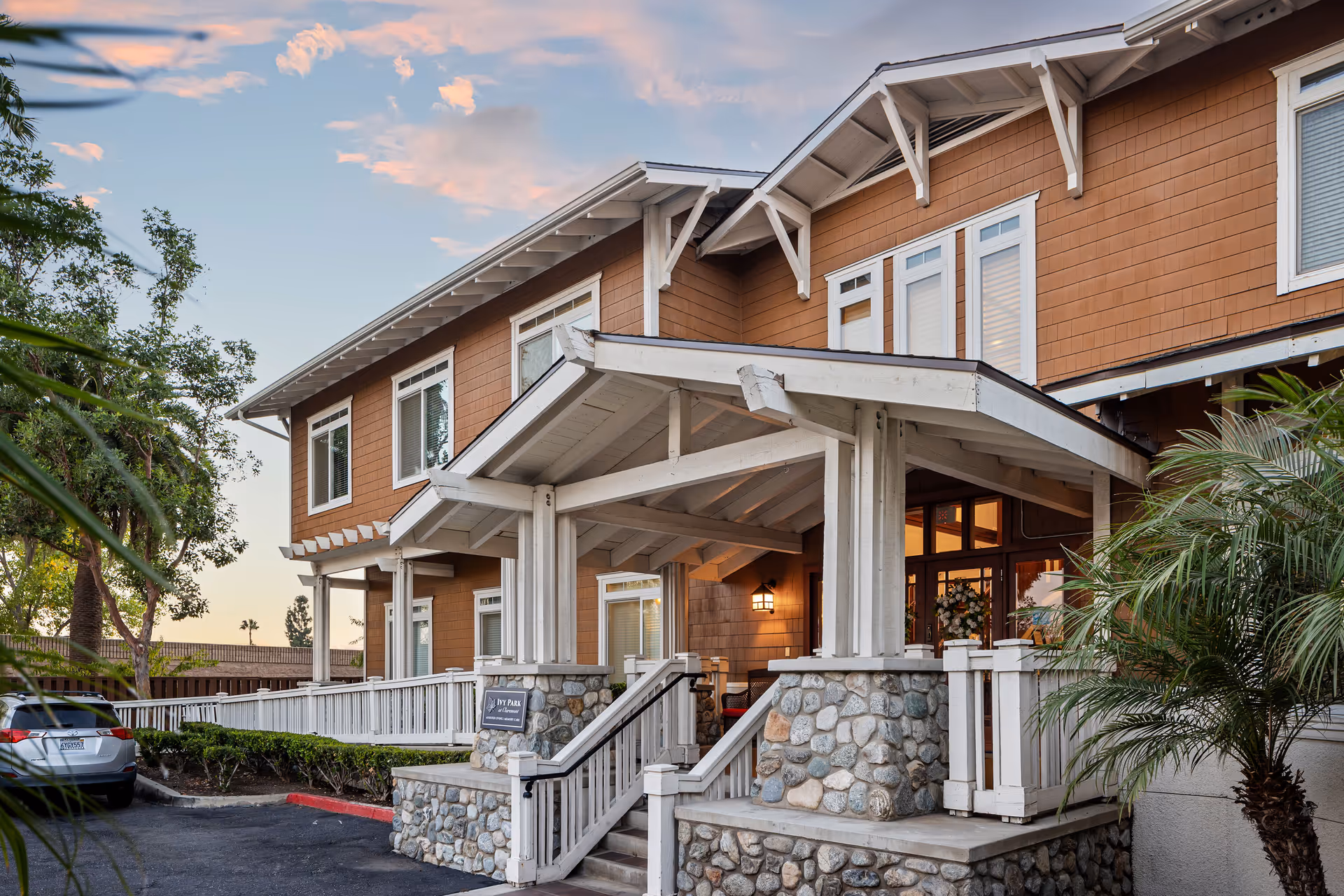 Exterior view of a two-story senior living facility building with brown siding and white trim. The entrance features a covered porch with stone pillars and white railings. There are trees and plants around the building, and a parked car is visible on the left side. The sky is partly cloudy with a warm light suggesting early evening.