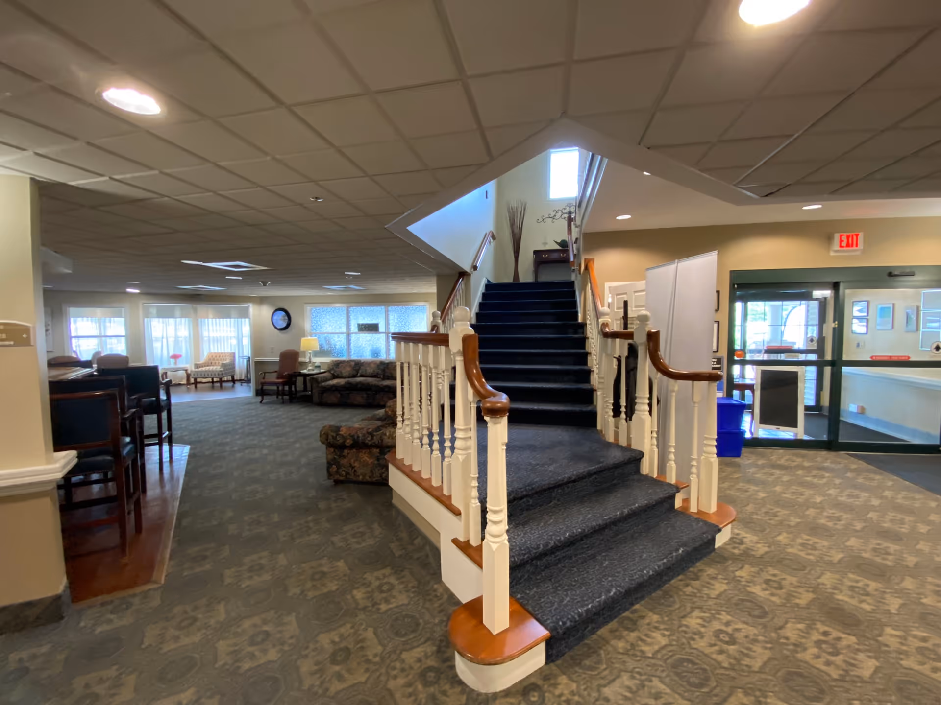 Interior view of a senior living facility lobby area with a carpeted staircase in the center, upholstered chairs and sofas arranged in seating areas, large windows letting in natural light, and an exit door to the right.