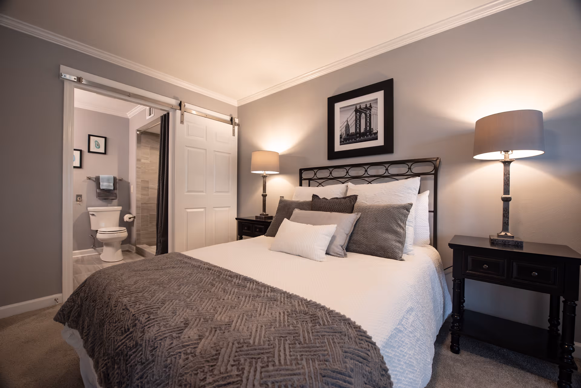 A cozy bedroom with a neatly made bed featuring multiple pillows and a textured gray blanket. On either side of the bed are dark wooden nightstands with matching lamps. A framed black and white photo hangs above the bed. To the left, a sliding barn door is partially open, revealing a bathroom with a toilet and a shower.