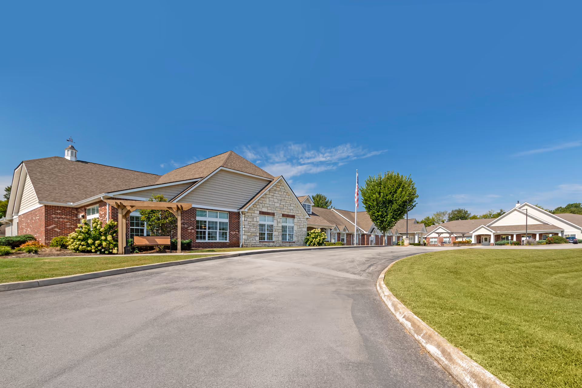 Exterior view of a senior living facility building with a curved driveway, well-maintained lawn, and clear blue sky. The building features brick and stone facades, multiple windows, a small pergola with a bench, and an American flag on a flagpole near the entrance.