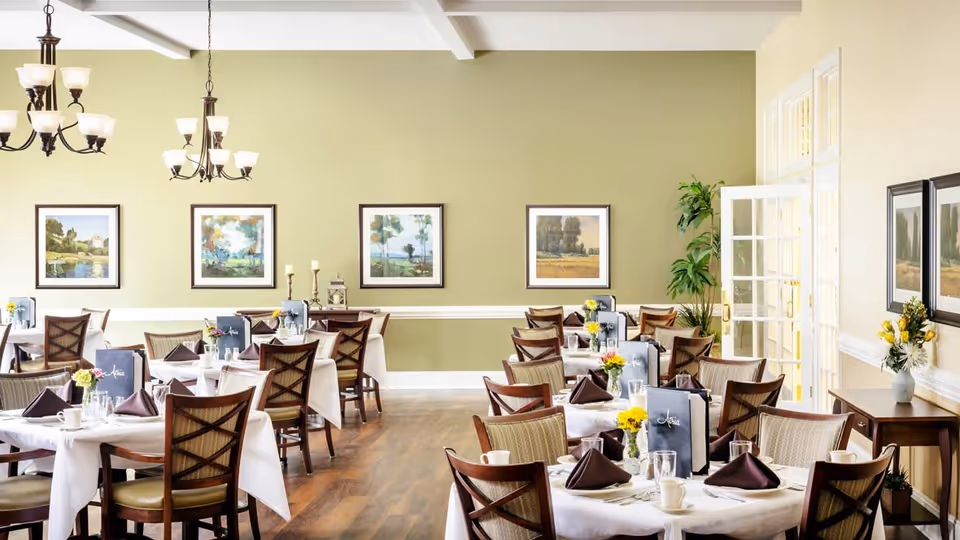 A bright and elegant dining room with multiple tables covered in white tablecloths, each set with brown napkins, menus, cups, and small flower arrangements. The room features wooden chairs, light green walls adorned with framed landscape paintings, two chandeliers hanging from the ceiling, and a wooden side table with a vase of flowers near a glass-paneled door.