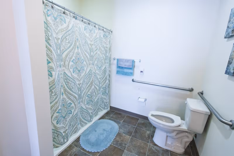 A clean bathroom with a white toilet, grab bars on the walls, a patterned shower curtain in shades of blue and green, a blue bath mat on the tiled floor, and two towels hanging on a towel rack.