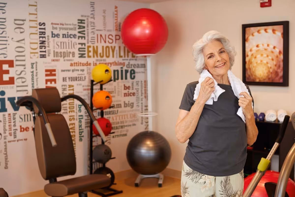 An elderly woman with white hair stands in a fitness room, smiling and holding a white towel around her neck. The room has exercise equipment including a weight machine, colorful exercise balls, and a large silver stability ball. The wall behind her features motivational words and phrases in various fonts and colors. A framed abstract artwork is visible on the wall.