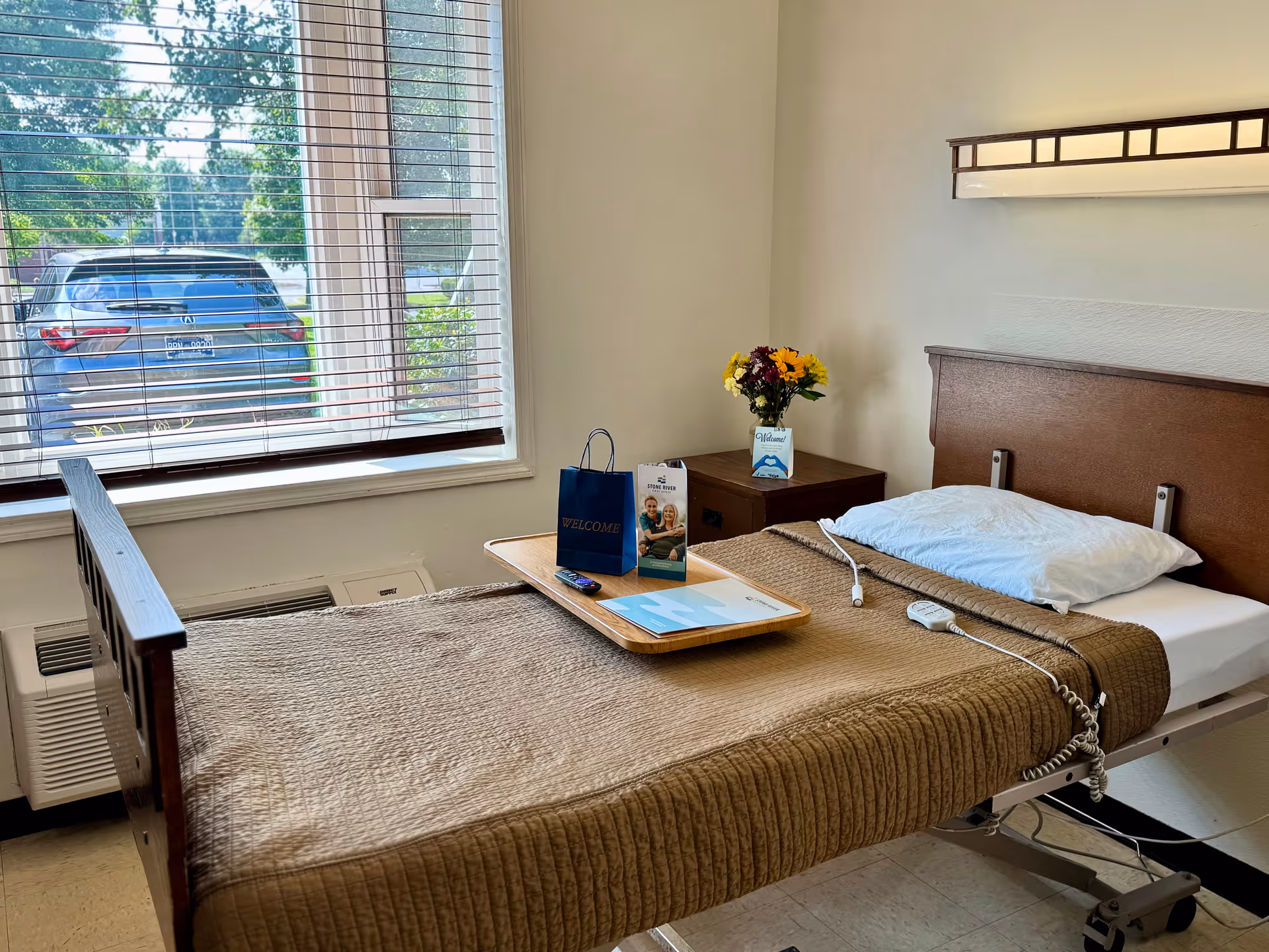 A single hospital-style bed with a brown quilted blanket and a white pillow in a small room. A wooden tray on the bed holds a welcome bag, a brochure, and a remote control. Next to the bed is a wooden nightstand with a vase of colorful flowers and a welcome card. A large window with blinds shows a parked car and greenery outside.