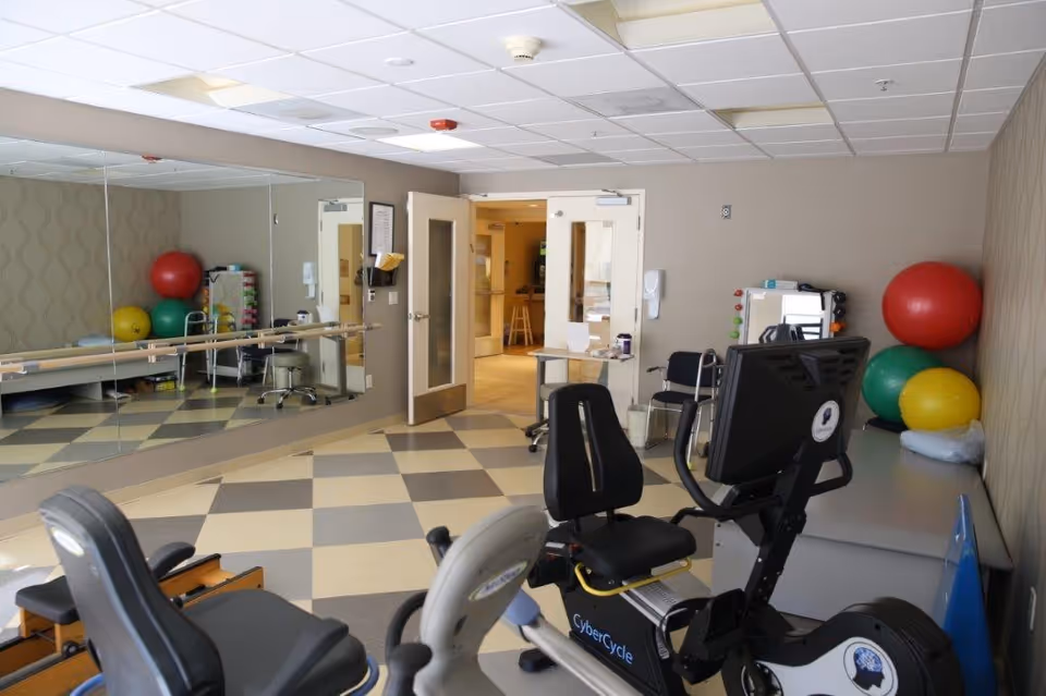 A physical therapy or exercise room with exercise equipment including a CyberCycle recumbent bike, large colorful exercise balls stacked in the corner, a wall-mounted ballet barre, and a large mirror covering one wall. The room has checkered flooring and beige walls, with double doors leading to another area.