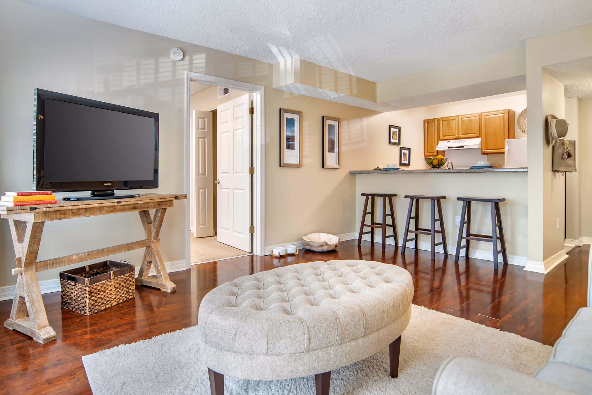 Spacious living room with a round tufted ottoman, TV on a wooden console, and a kitchen breakfast bar with three stools.