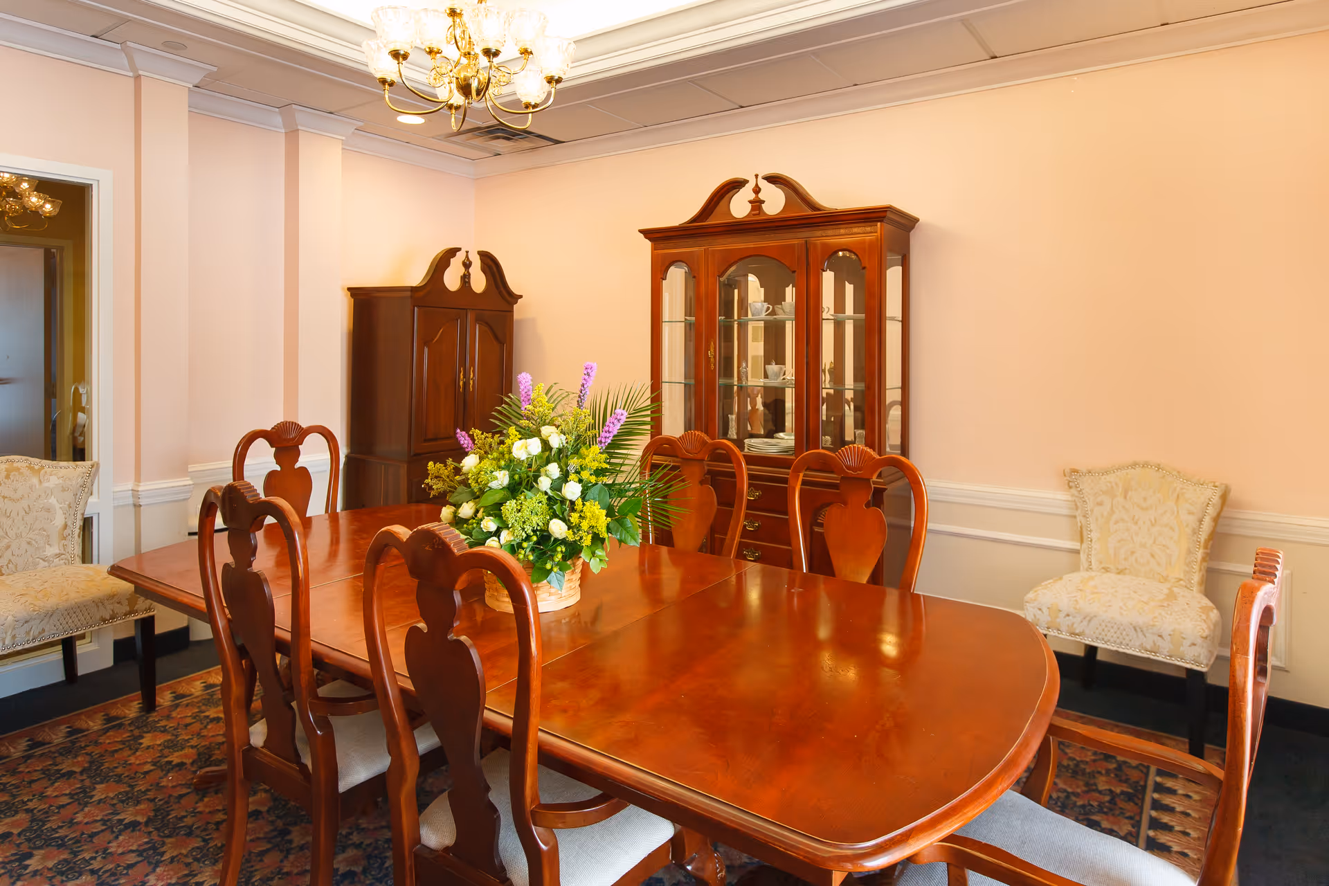 A traditional dining room with a polished wooden dining table surrounded by six matching chairs. A floral centerpiece with green and purple flowers is placed on the table. The room features a wooden china cabinet with glass doors displaying dishes, a wooden armoire, two upholstered accent chairs, a patterned rug, and a chandelier hanging from the ceiling. The walls are painted a soft peach color with white wainscoting.