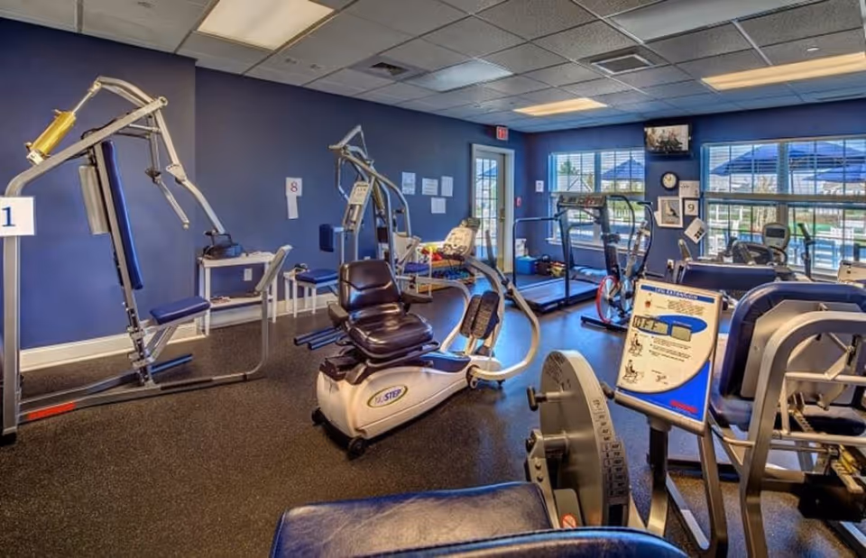 Interior view of a fitness room with various exercise equipment including weight machines, a recumbent bike, and treadmills. The room has blue walls, large windows with views of an outdoor pool area, and a TV mounted on the wall.