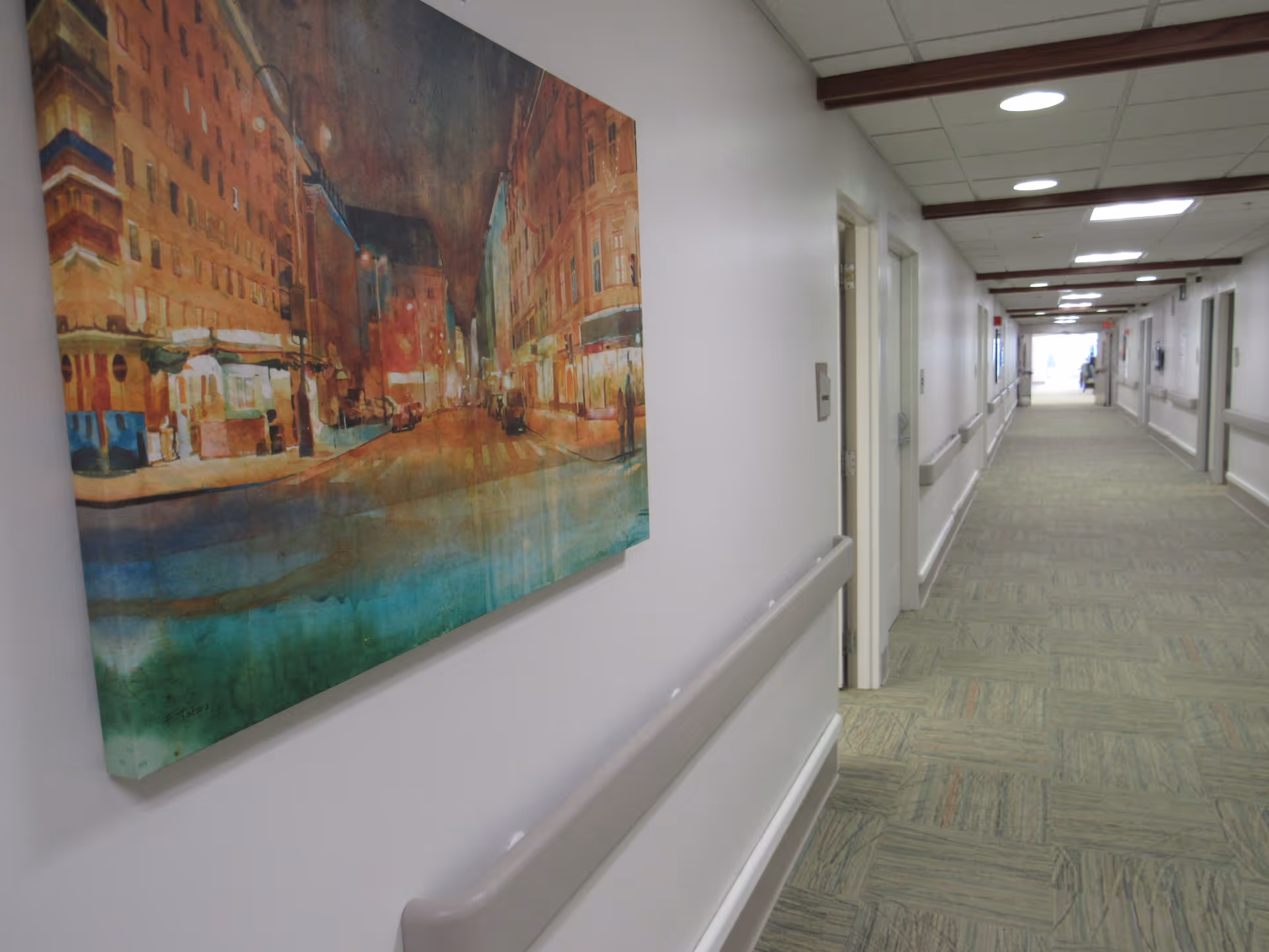 A long, well-lit hallway in a nursing and rehab center with handrails along the walls and a large colorful painting of a city street scene hanging on the left wall.