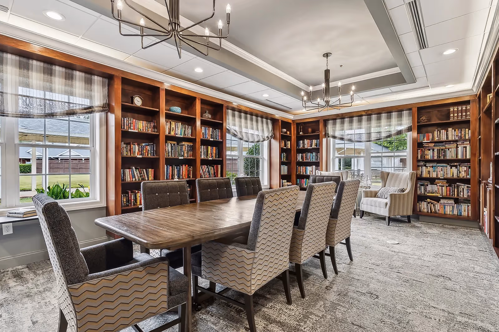 Well-lit library-style common room with a long wooden table surrounded by upholstered chairs and built-in bookshelves.